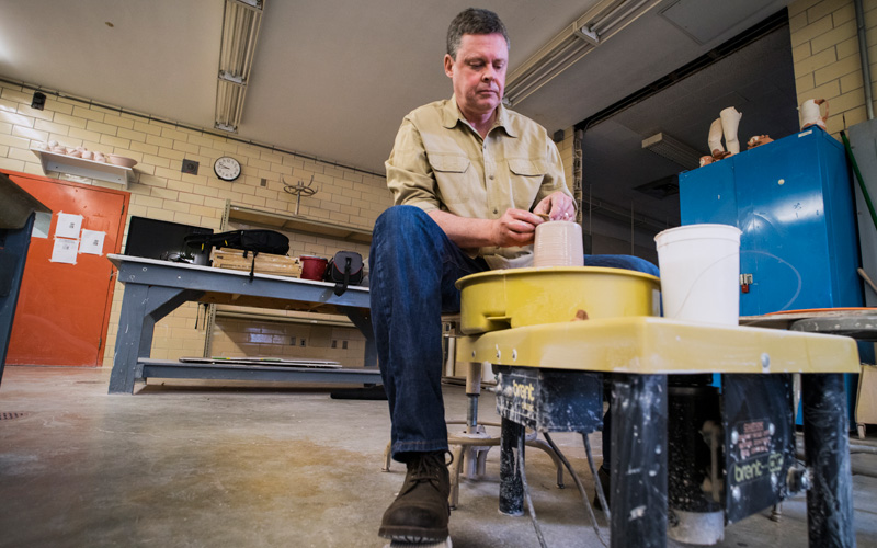 A man sitting at a potter's wheel shaping a case with his hands in a studio